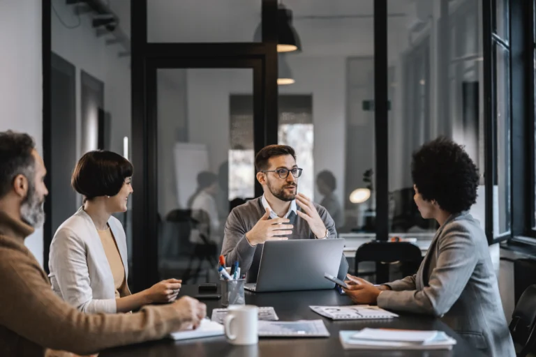 A professional business meeting in a modern office, featuring an experienced male lender with glasses speaking collaboratively and gesturing with his hands to a group of colleagues or clients seated around a conference table.
